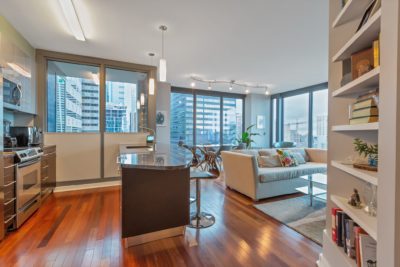 Modern open-concept apartment with a kitchen and living room. It features wood floors, a kitchen with stainless steel appliances, a cozy sofa, bookshelves, and large windows offering a city view. Airy and well-lit with contemporary decor.