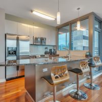 A modern kitchen with stainless steel appliances, including a fridge and stove. There’s a granite-topped island with three stylish wooden bar stools. Large windows offer a city view, and light wood flooring adds warmth to the space.