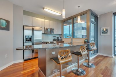 A modern kitchen with stainless steel appliances, including a fridge and stove. There’s a granite-topped island with three stylish wooden bar stools. Large windows offer a city view, and light wood flooring adds warmth to the space.