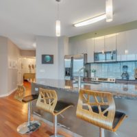 Modern kitchen with a sleek marble island, three wooden bar stools, and contemporary hanging lights. Stainless steel appliances and a blue-tiled backsplash enhance the space. Built-in shelves display books and decor beside the entrance to a hallway.