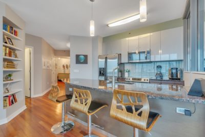 Modern kitchen with a sleek marble island, three wooden bar stools, and contemporary hanging lights. Stainless steel appliances and a blue-tiled backsplash enhance the space. Built-in shelves display books and decor beside the entrance to a hallway.
