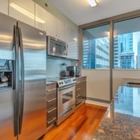 Modern kitchen with stainless steel appliances, including a fridge, oven, and microwave. Sleek countertops and wooden floors. Large window with a view of city skyscrapers.