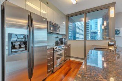 Modern kitchen with stainless steel appliances, including a fridge, oven, and microwave. Sleek countertops and wooden floors. Large window with a view of city skyscrapers.
