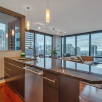 A modern apartment interior with a spacious kitchen featuring a dark granite island and stainless steel sink. Floor-to-ceiling windows offer a panoramic view of a cityscape with tall buildings and a cloudy sky. Cozy seating area visible in the background.