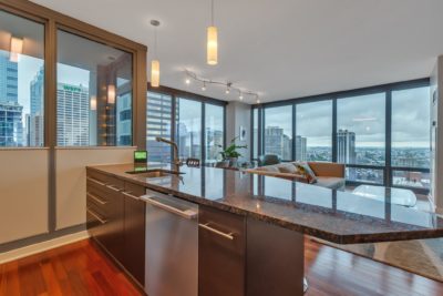 A modern apartment interior with a spacious kitchen featuring a dark granite island and stainless steel sink. Floor-to-ceiling windows offer a panoramic view of a cityscape with tall buildings and a cloudy sky. Cozy seating area visible in the background.