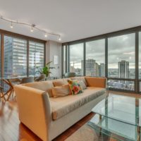 A modern living room with a beige sofa, glass coffee table, and wooden flooring. Large windows reveal a city skyline, and a dining table with chairs sits by the windows. Track lighting and a potted plant add to the decor.