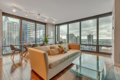 A modern living room with a beige sofa, glass coffee table, and wooden flooring. Large windows reveal a city skyline, and a dining table with chairs sits by the windows. Track lighting and a potted plant add to the decor.