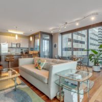 Modern living room with light wood floors, a beige couch, and glass coffee tables. The open kitchen features stainless steel appliances and bar seating. Large windows offer a city view, and a potted plant adds greenery. Contemporary track lighting is overhead.
