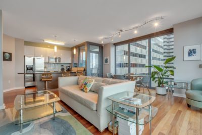 Modern living room with light wood floors, a beige couch, and glass coffee tables. The open kitchen features stainless steel appliances and bar seating. Large windows offer a city view, and a potted plant adds greenery. Contemporary track lighting is overhead.