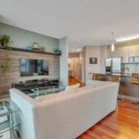A modern living room with a white couch facing a wall-mounted TV. The open-plan space includes a kitchen area with bar stools and stainless steel appliances. Wooden flooring and light gray walls create a contemporary and inviting atmosphere.