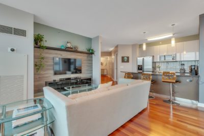 A modern living room with a white couch facing a wall-mounted TV. The open-plan space includes a kitchen area with bar stools and stainless steel appliances. Wooden flooring and light gray walls create a contemporary and inviting atmosphere.