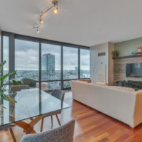 A modern living room with large windows offering a city view. A beige couch faces a wall-mounted TV on a wood-paneled wall. The room features a glass dining table with gray chairs and a large potted plant, and has wooden flooring.