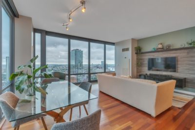 A modern living room with large windows offering a city view. A beige couch faces a wall-mounted TV on a wood-paneled wall. The room features a glass dining table with gray chairs and a large potted plant, and has wooden flooring.