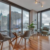 A modern dining area in a high-rise apartment with large windows showcasing a city skyline. The room features a glass table, four gray chairs, a potted plant, wooden floors, and a green armchair, set against a cloudy sky backdrop.