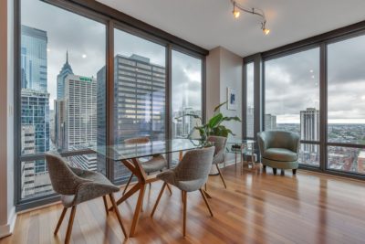 A modern dining area in a high-rise apartment with large windows showcasing a city skyline. The room features a glass table, four gray chairs, a potted plant, wooden floors, and a green armchair, set against a cloudy sky backdrop.