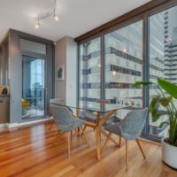 A modern dining area in an apartment with large floor-to-ceiling windows showing a city view. The space features a glass table with four gray chairs on wooden flooring. A potted plant and kitchen area with barstools are visible in the background.