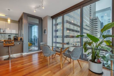 A modern dining area in an apartment with large floor-to-ceiling windows showing a city view. The space features a glass table with four gray chairs on wooden flooring. A potted plant and kitchen area with barstools are visible in the background.