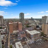 Aerial view of a cityscape featuring tall buildings, parking lots, and urban streets. The skyline is under a clear blue sky with scattered clouds. The architecture includes a mix of modern and older structures.