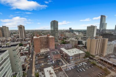 Aerial view of a cityscape featuring tall buildings, parking lots, and urban streets. The skyline is under a clear blue sky with scattered clouds. The architecture includes a mix of modern and older structures.