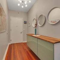 A modern hallway with a wooden floor and a wall-mounted cabinet with a wooden top. Three round mirrors and a tall vase with branches sit on the cabinet. The walls are decorated with framed art. A chandelier hangs from the ceiling near the white door.