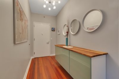 A modern hallway with a wooden floor and a wall-mounted cabinet with a wooden top. Three round mirrors and a tall vase with branches sit on the cabinet. The walls are decorated with framed art. A chandelier hangs from the ceiling near the white door.