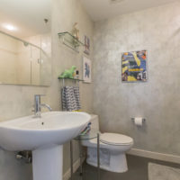 A modern bathroom with a white pedestal sink, a toilet, and a glass-enclosed shower. The walls feature framed art, and there are decorative items on a shelf. A small carpet lies on the floor in front of the sink.