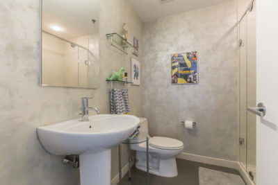 A modern bathroom with a white pedestal sink, a toilet, and a glass-enclosed shower. The walls feature framed art, and there are decorative items on a shelf. A small carpet lies on the floor in front of the sink.