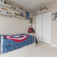 A neatly organized bedroom features a bed with a Captain America-themed comforter. Shelves on the light gray walls display toys and decorations. A closet with white sliding doors is on the right, and several hats are hung nearby.