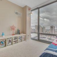 A modern kids' room with superhero-themed storage bins and bedding. A lava lamp and toys sit on a low shelf. Large windows offer a view of skyscrapers under a cloudy sky. Neutral walls and carpet provide a clean, bright atmosphere.