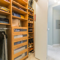 A well-organized walk-in closet with wooden shelves and drawers. Clothes and handbags are neatly stored, and shoes are lined up on racks. The closet opens into a carpeted hallway with a white door.