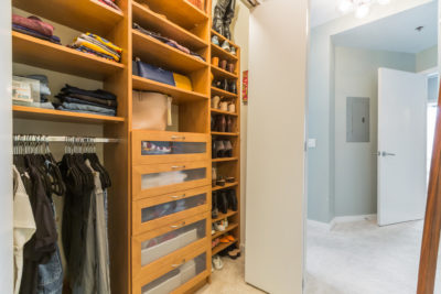 A well-organized walk-in closet with wooden shelves and drawers. Clothes and handbags are neatly stored, and shoes are lined up on racks. The closet opens into a carpeted hallway with a white door.