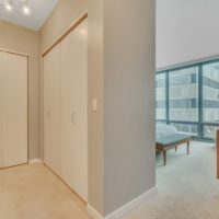 A modern bedroom with beige carpeting features sliding closet doors, a wooden dresser, and a wide window offering a view of nearby buildings. A bed with pillows is positioned near the window.