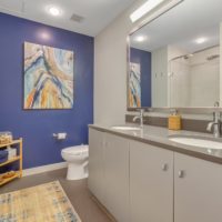 A modern bathroom with dual sinks, a large mirror, and a blue accent wall featuring abstract art. A light wood shelf holds towels and a rug lies on the floor near the shower. The overall decor is contemporary and clean.