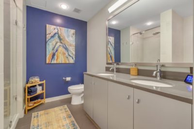 A modern bathroom with dual sinks, a large mirror, and a blue accent wall featuring abstract art. A light wood shelf holds towels and a rug lies on the floor near the shower. The overall decor is contemporary and clean.