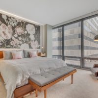 A modern bedroom with a large bed adorned with gray bedding and pink pillows. There's a floral mural above the bed, a floor lamp, and tall glass windows offering a view of city buildings. A bench is at the foot of the bed.