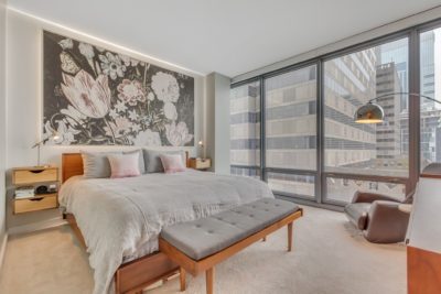 A modern bedroom with a large bed adorned with gray bedding and pink pillows. There's a floral mural above the bed, a floor lamp, and tall glass windows offering a view of city buildings. A bench is at the foot of the bed.