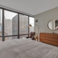 A modern bedroom with a large bed, a wooden dresser with a round mirror, and a leather chair by a floor lamp. Tall windows offer a cityscape view of skyscrapers. Neutral tones create a calm atmosphere.