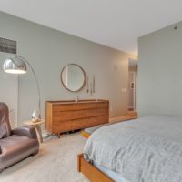 A modern bedroom with a beige carpet, featuring a gray bed and a wooden dresser with a round mirror above it. A brown leather armchair and a tall floor lamp are near a large window, allowing natural light to fill the space.