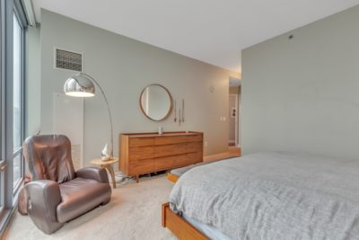 A modern bedroom with a beige carpet, featuring a gray bed and a wooden dresser with a round mirror above it. A brown leather armchair and a tall floor lamp are near a large window, allowing natural light to fill the space.
