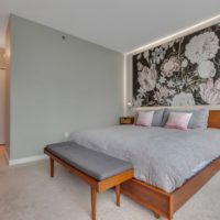 A modern bedroom with a large bed featuring gray bedding and pink pillows. A floral mural decorates the wall behind the bed. A wooden bench and nightstands flank the bed. Soft lighting illuminates the room, creating a cozy atmosphere.