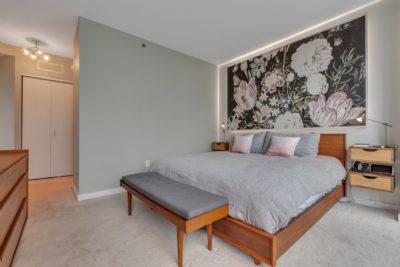 A modern bedroom with a large bed featuring gray bedding and pink pillows. A floral mural decorates the wall behind the bed. A wooden bench and nightstands flank the bed. Soft lighting illuminates the room, creating a cozy atmosphere.