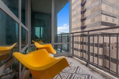 Balcony view with two modern yellow chairs and a patterned rug against a backdrop of tall buildings. The sky is clear and blue, visible through the glass panels on the left.