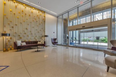 A modern hotel lobby features a large glass entrance, a polished floor, and a gold-accented wall. It is furnished with a brown sofa, chairs, and sleek lighting, creating an inviting atmosphere.