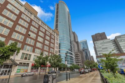 A city street with modern high-rise buildings on a clear day. The foreground features a curved glass skyscraper, cars parked along the road, and a few pedestrians. The sky is bright blue with a few clouds scattered.