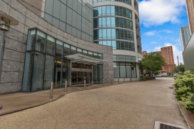 Modern building entrance with large glass windows and a canopy. Sidewalk features textured paving and is flanked by greenery. Tall structures and clear blue sky in the background.